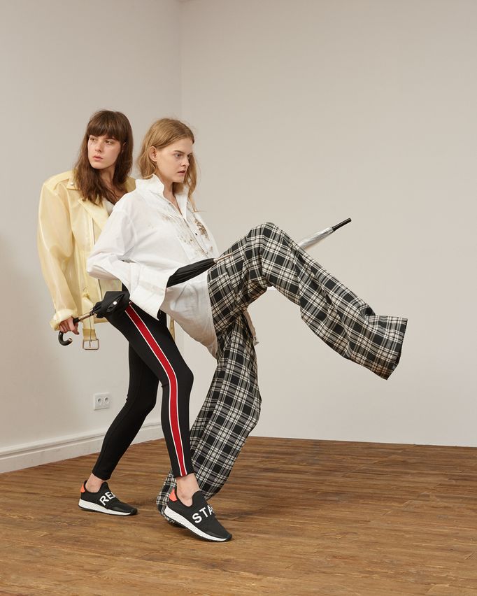 Two fashion models posing indoors: one wearing a loose yellow satin shirt with black leggings featuring red side stripes, the other in an oversized white shirt styled with wide plaid trousers. Both wear slip-on sneakers with bold text prints, creating a mix of sporty and avant-garde style.